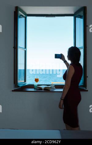 Spanien, Kanarische Inseln, La Palma, junge und hübsche Frau, die den Horizont von einem Hotelzimmer aus, durch ein Fenster, am Meer fotografiert Stockfoto