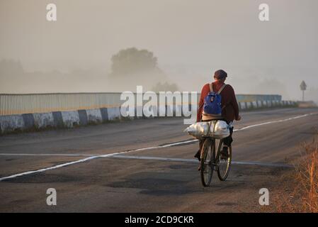 Großvater in alten Kleidern mit einer großen Tasche fährt an einem nebligen Morgen, dem Thema Armut, mit dem Fahrrad auf einer asphaltierten Straße. Stockfoto