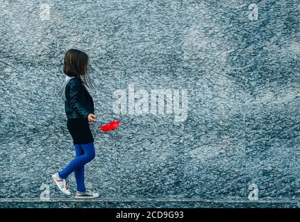 Kleine Stadt stilvolle Mädchen in lässigen Herbst Kleidung zu Fuß entlang graue Granitwand mit rotem Wind Spinner. Leerzeichen für Text. Stockfoto