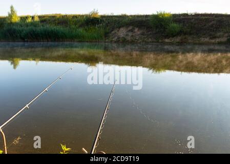 Zwei Angelruten stehen am sandigen Ufer eines Fischteich, beleuchtet von der Morgensonne. Stockfoto