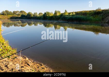 Zwei Angelruten stehen am sandigen Ufer eines Fischteich, beleuchtet von der Morgensonne. Stockfoto