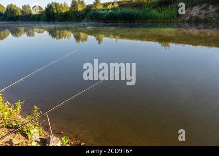 Zwei Angelruten stehen am sandigen Ufer eines Fischteich, beleuchtet von der Morgensonne. Stockfoto