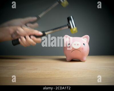 Eine Hand, die einen Hammer hält, der über einem rosa, traurigen Sparschwein angehoben wird, mit einem schockierten und ängstlichen Gesichtsausdruck. Reflektierende Oberfläche und leicht g Stockfoto