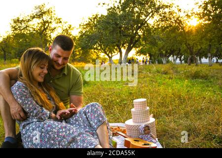 Glückliches junges Paar entspannend und mit Picknick im Park. Friedliche süße Pärchen genießen Abendessen im Park. Mann und Frau sitzen auf Gras und umarmen. Stockfoto