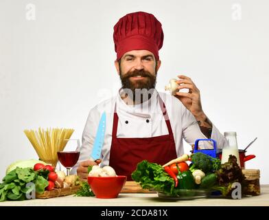 Konzept der Restaurantküche. Der Küchenchef bereitet das Essen zu. Kochen mit Lächeln in burgunderroten Uniform sitzt am Küchentisch mit Gemüse und Geschirr. Mann mit Bart hält Pilz und Messer auf weißem Hintergrund Stockfoto