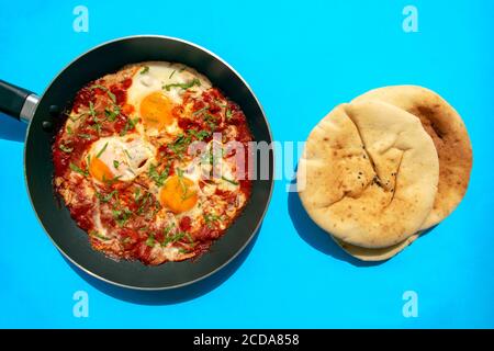 Shakshuka - pochierte Eier in Tomatensauce, Zwiebel, Pfeffer und Gewürze in eiserner Pfanne mit Pita Brot auf blauem hellen Hintergrund. Berühmte traditionelle arabische an Stockfoto