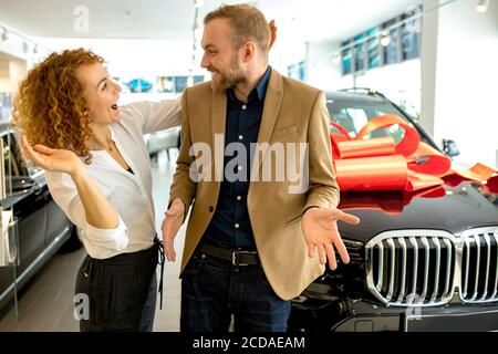 Schöne kaukasische Dame überraschen Ehemann in Händler. Frau geben neues Auto in roten Bogen zu ihrem Mann eingewickelt, Geburtstagsgeschenk. Sie öffnet Mann Stockfoto