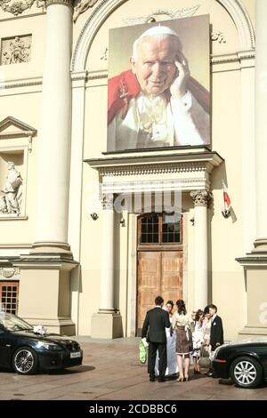 WARSCHAU, POLEN - 12. JULI 2009: Frisch verheiratete junge Menschen stehen vor einer Kirche mit einem riesigen Bild von Johannes Paul II., oder Karol Wojtyla, der o Stockfoto