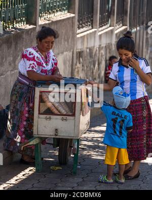Eine junge Tzutujil Maya Frau kauft ein Eis von einem Händler auf der Straße in San Pablo la Laguna, Guatemala. Der Verkäufer trägt das traditionelle Kleid Stockfoto