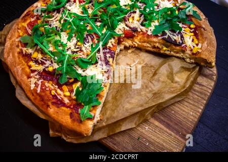 Hausgemachte Pizza mit Rucola, Parmesankäse und roter Sauce mit einem Stück auf Pergament dunklen Hintergrund, close-up Stockfoto