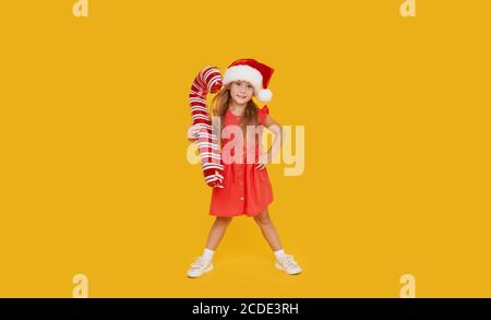 Ein niedliches kleines Mädchen Kind in einem ausgefallenen Kleid und einem Weihnachtsmann Hut hält eine aufblasbare Form eines Zuckerrohrs auf einem gelben Hintergrund. 2021 Neujahr con Stockfoto