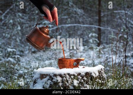 Ein Mann gießt aromatischen Kaffee in eine Holzschale im Wald. Die Tasse steht auf einem alten Stumpf. Der Hintergrund des Waldes ist verschwommen. Stockfoto