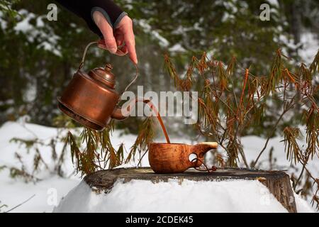 Ein Mann gießt aromatischen Kaffee in eine Holzschale im Wald. Die Tasse steht auf einem alten Stumpf. Der Hintergrund des Waldes ist verschwommen. Stockfoto