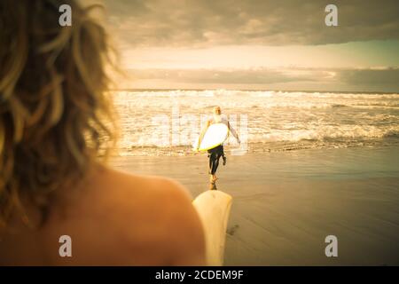 Glückliche Familie, die Spaß beim Surfen im Sonnenaufgang hat. Der Sohn des Surfers wartet am Ufer auf seinen Vater. Sportlicher People Lifestyle und Extremsportkonzept Stockfoto