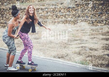 Junge Mutter übt auf Skateboard in der ländlichen Straße. Tochter hilft Mama auf Skateboard. Mama lernen, Skateboard zu fahren, wie Tochter lehrt sie Stockfoto