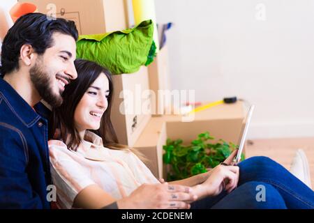 Glückliches junges Paar mit Tablet für neues Haus Projekt. Mann und Frau sitzen in der Nähe von Kartons in neuen Grundstückshaus. Mieten, Kaufen, Lifestyle und Relatio Stockfoto