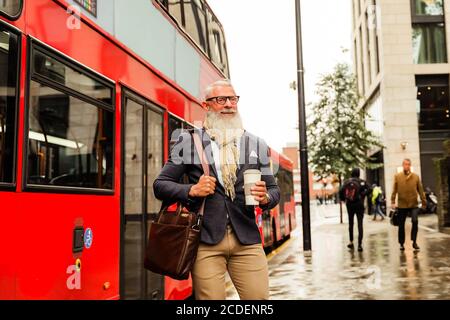 Glücklicher Geschäftsmann trinkt Kaffee und verlässt die Arbeit. Mann, der an einem regnerischen Tag mitten im Herbst läuft. Mann, der aus dem Bus kommt. Arbeit und fa Stockfoto