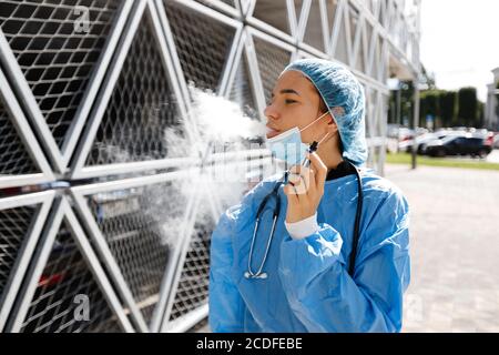Müde junge Arzt oder Krankenschwester auf Pause nach der Operation Rauchen elektronische Zigarette trägt chirurgische Gesichtsmaske, Kappe und Kleid in der Nähe des Krankenhauses Stockfoto