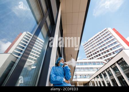 Weitwinkel: Junge Arzt oder Krankenschwester auf Pause raucht elektronische Zigarette trägt chirurgische Gesichtsmaske, Kappe und Kleid in der Nähe des Krankenhauses Stockfoto