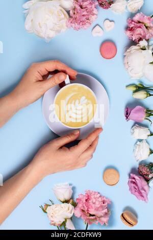 Festliches vertikales Foto mit blauem Hintergrund. Eine Tasse Latte Macchiato in Frauenhänden, umgeben von Rosen, Pfingstrosen, Makronen und Zuckerstücken i Stockfoto