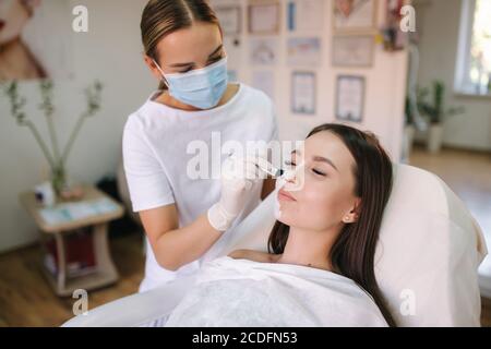 Junge Frau Kosmetologe gelten weißen Ton Maske auf Frau Gesicht. Schöne Brünette Frau im Schönheitssalon Stockfoto