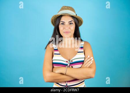 Junge schöne Frau trägt Badeanzug und Hut über isoliert blau Hintergrund gekreuzt Arme Blick auf die Kamera lächelnd Stockfoto