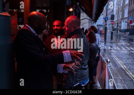 In einer regnerischen Nacht in Soho, schützen männliche Trinker unter einem Baldachin und einem warmen roten Licht vor dem Pub „Nellie Dean of Soho“ in der Dean Street, zu einer Zeit, als kürzlich wiedereröffnete Bars und Restaurants während der Coronavirus-Pandemie am 27. August 2020 verzweifelt nach Kundengeschäft suchen, In London, England. Stockfoto