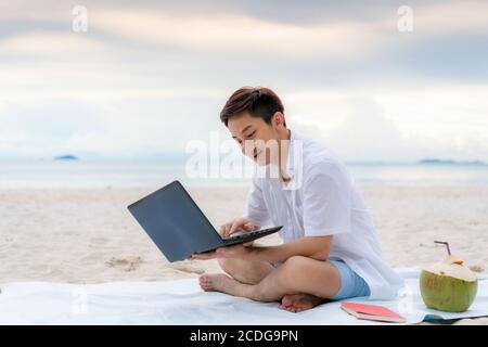 Junger asiatischer Mann, der während seiner Urlaubszeit mit einem Laptop draußen arbeitet, während er am schönen Strand sitzt. Sommer, Urlaub, Urlaub und glückliche Menschen ich Stockfoto