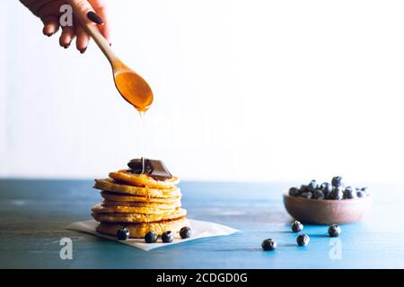 Eine anonyme Frau Hand gießt Sirup auf minimal suchen Gestapelte Pfannkuchen mit Heidelbeeren über blauem Holz Vintage Hintergrund mit Speicherplatz kopieren Stockfoto
