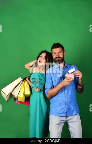 Liebespaar hält Einkaufstaschen auf grünem Hintergrund. Kerl mit Bart und Mädchen mit nachdenklichen Gesichtern tun Einkaufen. Mann mit Bart hält Kreditkarte und Sparschwein. Einkaufs- und Ausgabenkonzept. Stockfoto