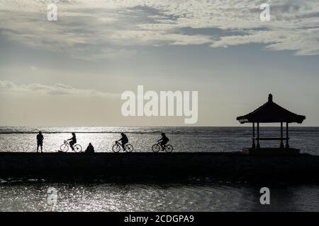 Genießen Sie die morgendliche Atmosphäre beim Radfahren am Strand von Bali mit Freunden. Gesunde Bewegung während der COVID-19 Pandemie. Pier mit balinesischen Gebäuden Stockfoto