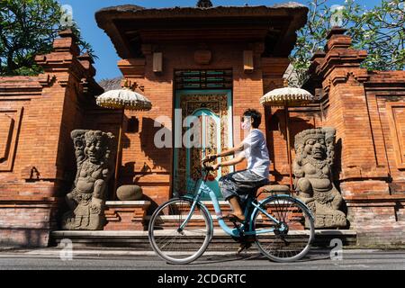 Eine Frau, die im Sommer auf Bali Fahrrad fährt. Traditionelle balinesische Türen aus roten Ziegeln sorgen für eine schöne und attraktive Kulisse Stockfoto