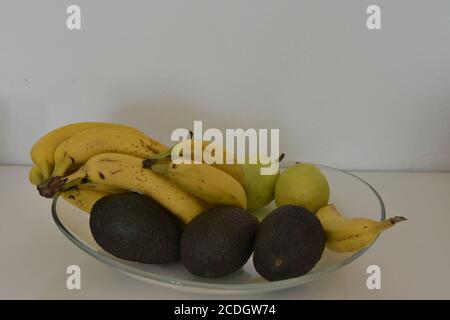 Stillleben der Anordnung von Bananen, Birnen und Avocados auf einer großen Glasablage. Die isolierten Früchte dienen sowohl als Dekoration als auch als Angebot von Snacks. Stockfoto