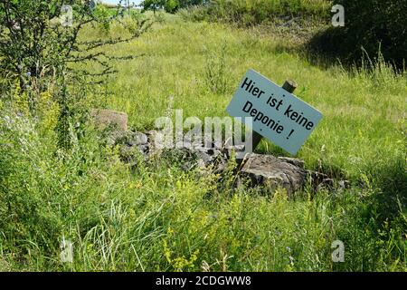 Warnschild in deutscher Sprache, hier ist kein Dumpingplatz, zwischen Steinen und Gras gelegen. Alarmtafel hat rechteckige Form, ist aus Holz. Stockfoto