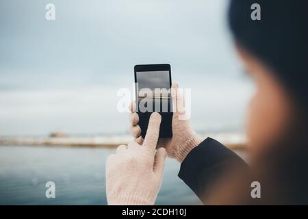 Weibliche Hände tragen warme Handschuhe halten Smartphone fotografieren Meer Am bewölkten Wintertag Stockfoto