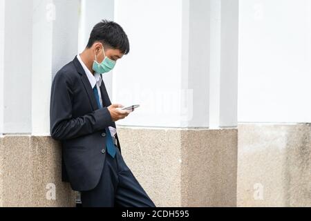 Ein hübscher junger asiatischer Geschäftsmann mit Smartphone. Asiatische junge attraktive Geschäftsmann trägt eine Gesichtsmaske stehen gegen die Wand. Stockfoto