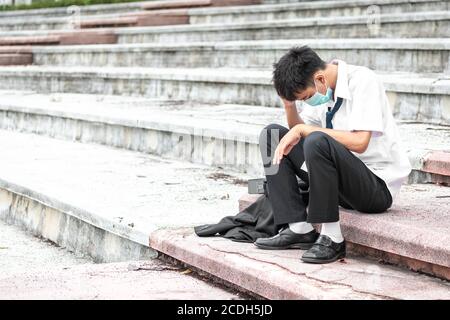 Ausfall arbeitslos gestresst jungen asiatischen Geschäftsmann trägt eine Gesichtsmaske, die Kopf mit Händen. Ein junger Geschäftsmann sitzt verzweifelt auf der Treppe Stockfoto
