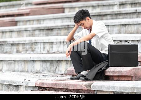 Ausfall arbeitslos gestresst junge asiatische Business-Mann über Kopf mit Händen. Ein junger Geschäftsmann sitzt verzweifelt auf der Treppe, weil niedrige economii Stockfoto