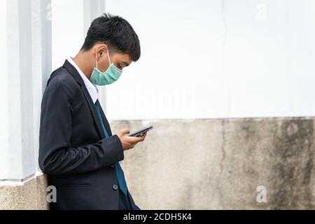 Ein hübscher junger asiatischer Geschäftsmann mit Smartphone. Asiatische junge attraktive Geschäftsmann trägt eine Gesichtsmaske lehnt an der Wand. Stockfoto