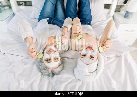 Draufsicht Porträt zeigt zwei Frauen, ältere hübsche Großmutter und junge lächelnde Enkelin, die zu Hause auf dem Bett liegen, mit Gesichtsmasken im Gesicht Stockfoto