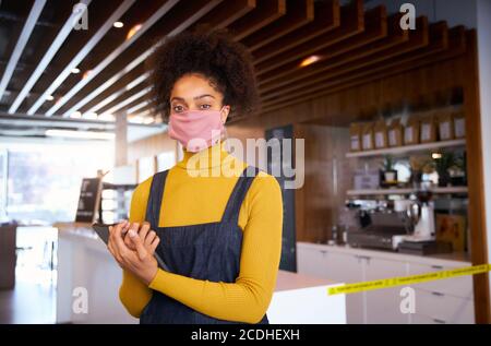 Weibliche afrikanische Café-Besitzer trägt Gesichtsmaske stehen vor der Theke mit digitalen Tablet. Stockfoto