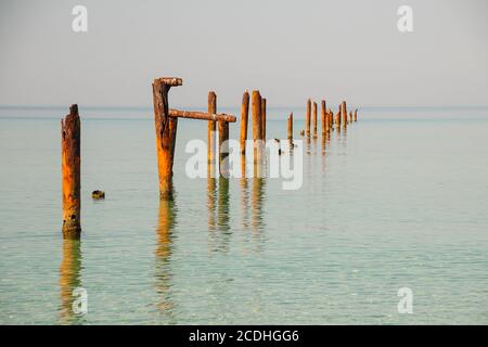 Seascape mit blauem ruhigem Wasser und Reihen von rostigen Rohren mit grünen Algen am Sommertag. Rostpfeifen, die vom alten Pier übrig geblieben sind. Perspektive, die zu führt Stockfoto