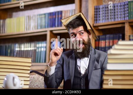 Lehrer mit Bart und geöffnetem Buch auf dem Kopf, wie ein Dach. Mad Professor Konzept. Mann im klassischen Anzug oder altmodischen Professor mit glücklichen Gesicht sitzt in der Bibliothek, Bücherregale auf dem Hintergrund, defokussiert. Stockfoto
