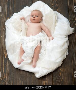 Kleinkind mit weißer Decke bedeckt. Kindheit und Göttliche Geschenk Konzept. Neugeborenes Kleinkind mit blauen Augen und interessiertes Gesicht auf Holzhintergrund. Baby liegt auf weißen weichen Bettdecke als Wolke oder Nest entworfen Stockfoto