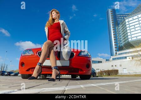 Schöne blonde Frau in einem roten Kleid posiert in einem Rotes Auto in der Stadt an einem sonnigen Tag Stockfoto