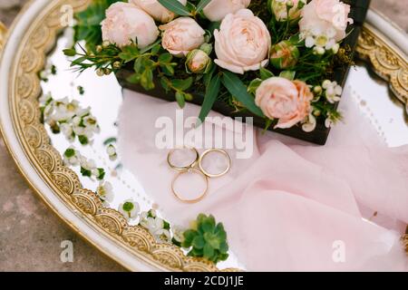 Zwei Eheringe für Braut und Bräutigam und der Verlobungsring der Braut auf einem Tablett mit Mustern und einem Strauß rosa Rosen. Stockfoto
