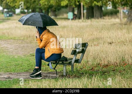 WIMBLEDON LONDON, GROSSBRITANNIEN - 28. AUGUST 2020 . Eine Frau, die auf einer Bank sitzt, hält einen Regenschirm von den Regenschauern auf Wimbledon Common. Kredit: amer ghazzal/Alamy Live Nachrichten Stockfoto