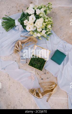 Zwei Eheringe und Brautaccessoires auf weißem Stoff mit Blumenstrauß und Sandalen mit Fersen. Stockfoto