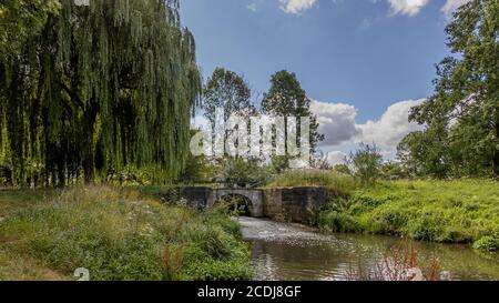 Keutelbeek Bach im Stadtpark Sittard umgeben von Bäumen und grüner Vegetation mit einer kleinen Brücke über den Damm und der Schleuse im Hintergrund, Stockfoto