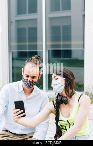 Nahaufnahme eines jungen Paares mit einer Gesichtsmaske auf einer Bank auf der Straße sitzen Spaß mit ihrem Handy. Stockfoto